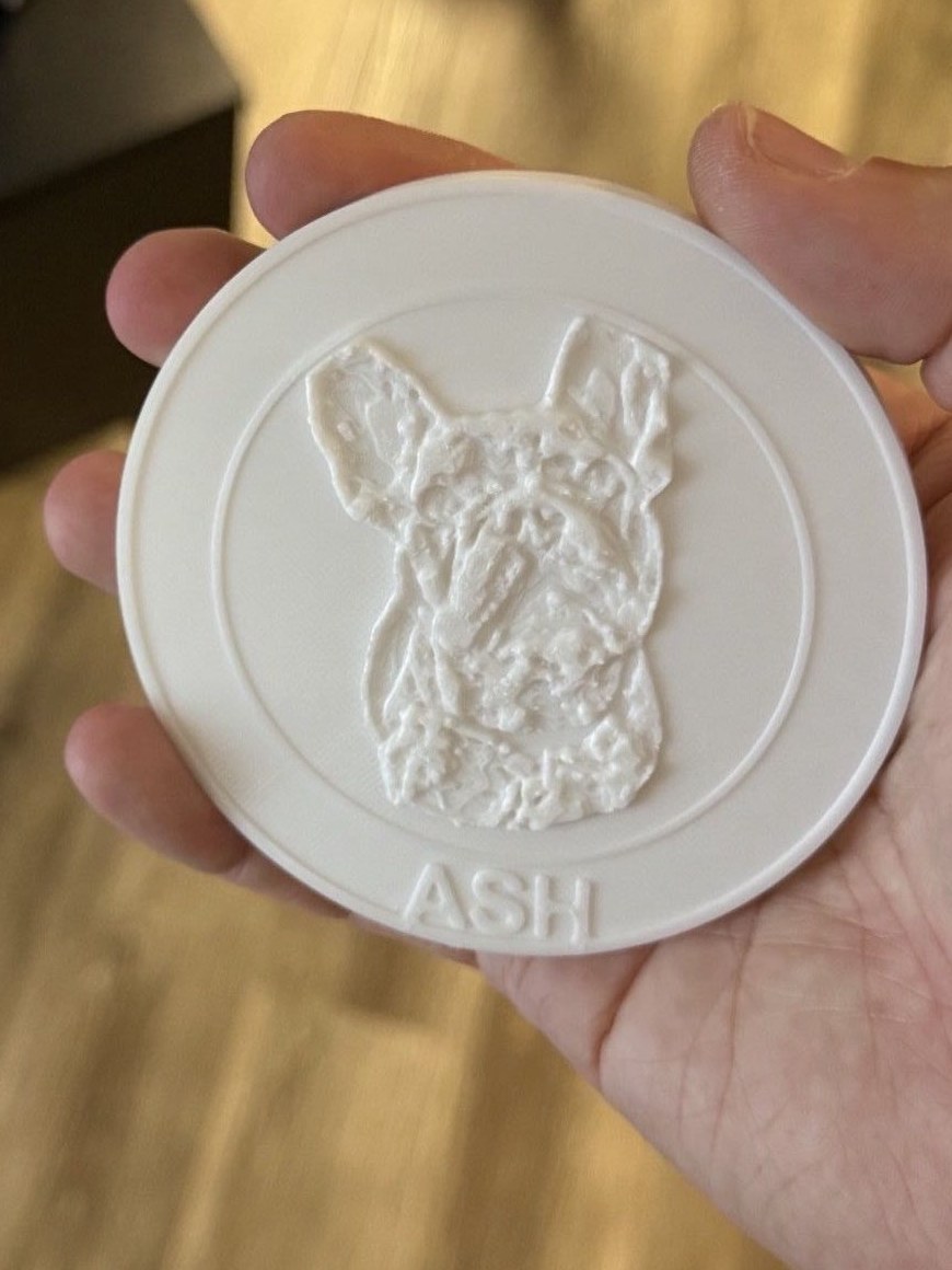 A white 3D-printed round coaster showing a relief portrait of a French bulldog and the name ASH along the bottom edge, held in a hand