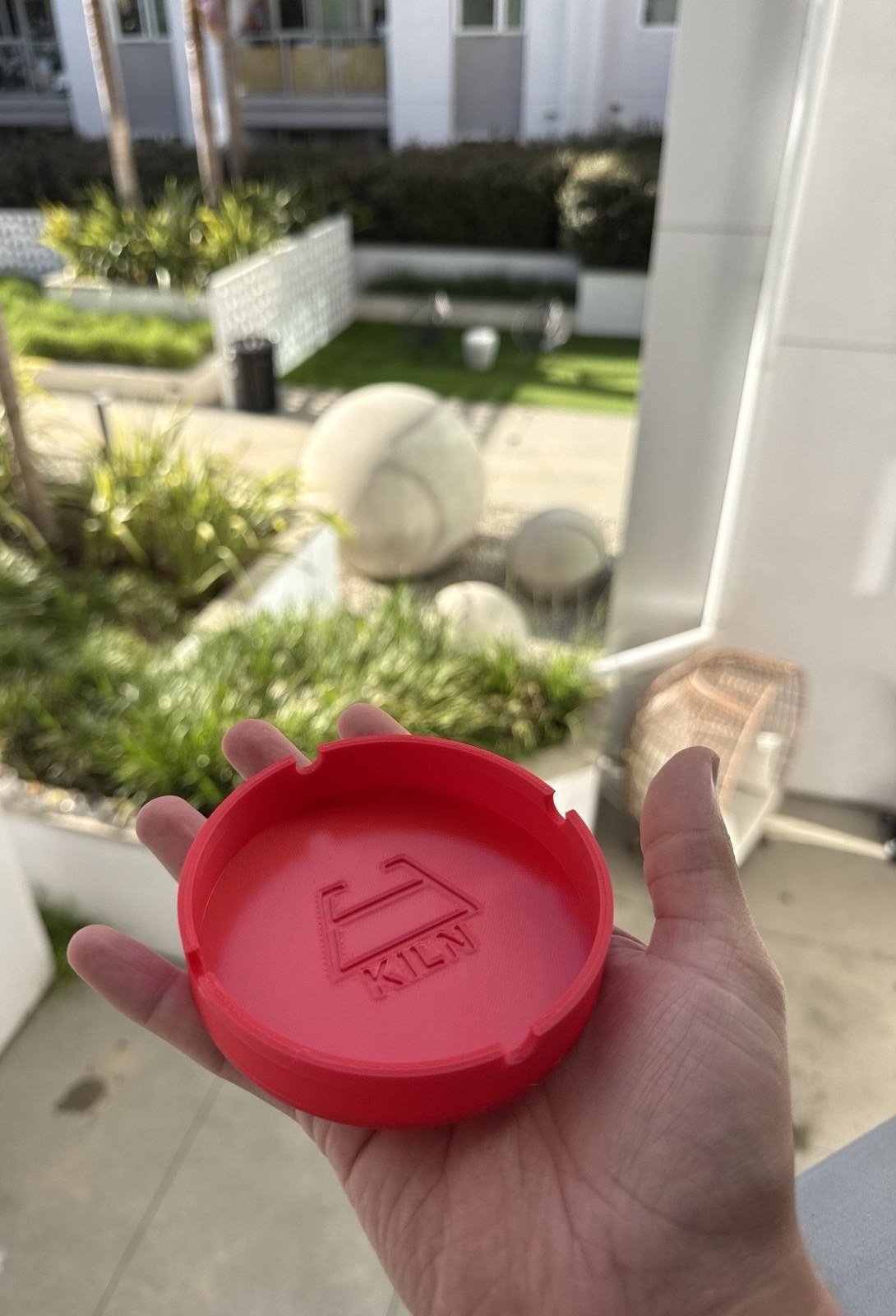 A red 3D-printed circular ashtray held in a hand, with the Kiln mark debossed into the inside